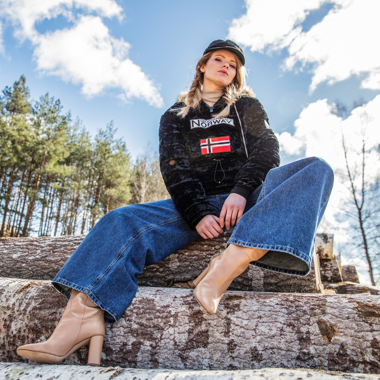Frau, die ein Geographical Norway Sweatshirt mit der norwegischen Flagge trägt, sitzt auf Baumstämmen mitten in der Natur. Ein lässiger und trendiger Look für moderne Abenteurerinnen