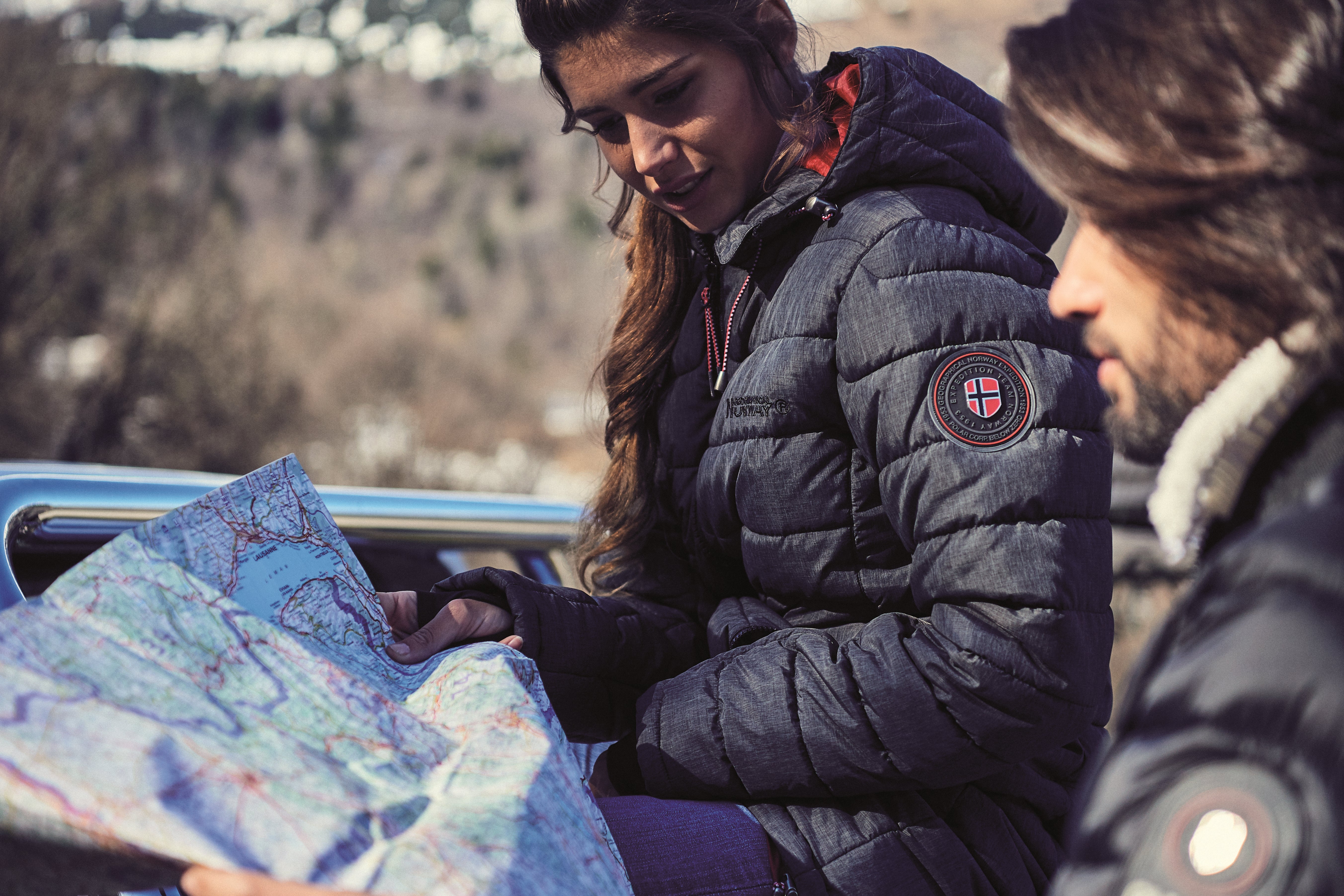 Femme portant une doudoune Geographical Norway, lisant une carte avec un compagnon en pleine nature. Un look parfait pour les explorateurs et les amateurs de voyages en extérieur
